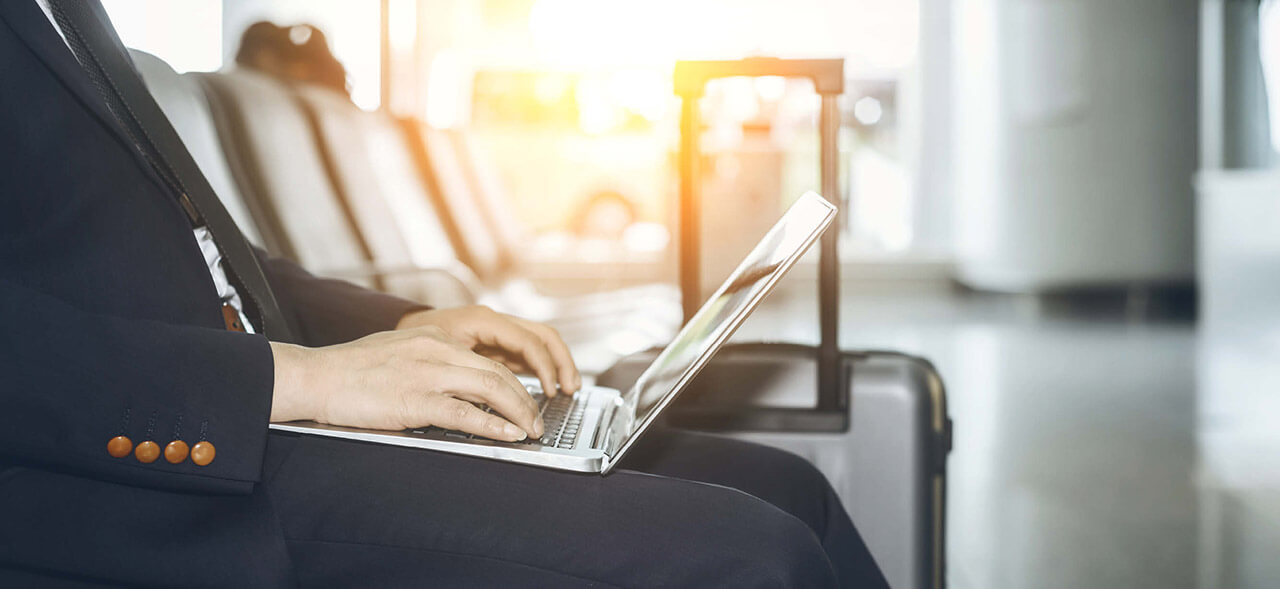 Image of a man in business suit using a laptop at the airport terminal with a suitcase seen next to him